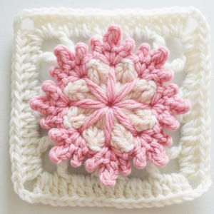 May include: A handmade crochet square with a central pink and cream floral motif. The flower is set within a white square frame with an openwork design. This piece is likely part of a larger craft project, such as a blanket or cushion.