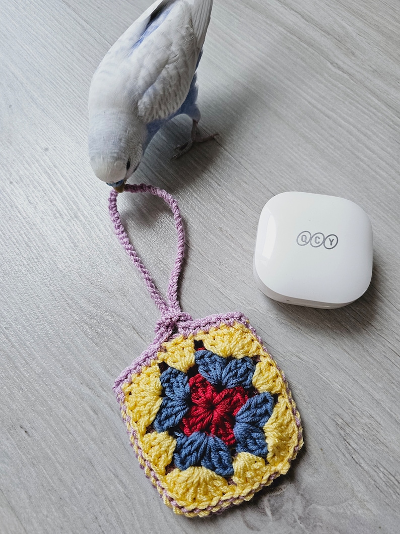 May include: A crocheted square with a purple border, yellow, blue, and red yarn. The square is hanging from a purple cord. A small white device with the letters QCY is in the background.