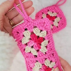 Op de afbeelding: Roze gehaakte telefoonhoes en bijpassende vierkante onderzetter. De hoes heeft een bloemenpatroon met witte en rode accenten. De hoes heeft een lus om te dragen. De onderzetter heeft een vergelijkbaar bloemenpatroon.