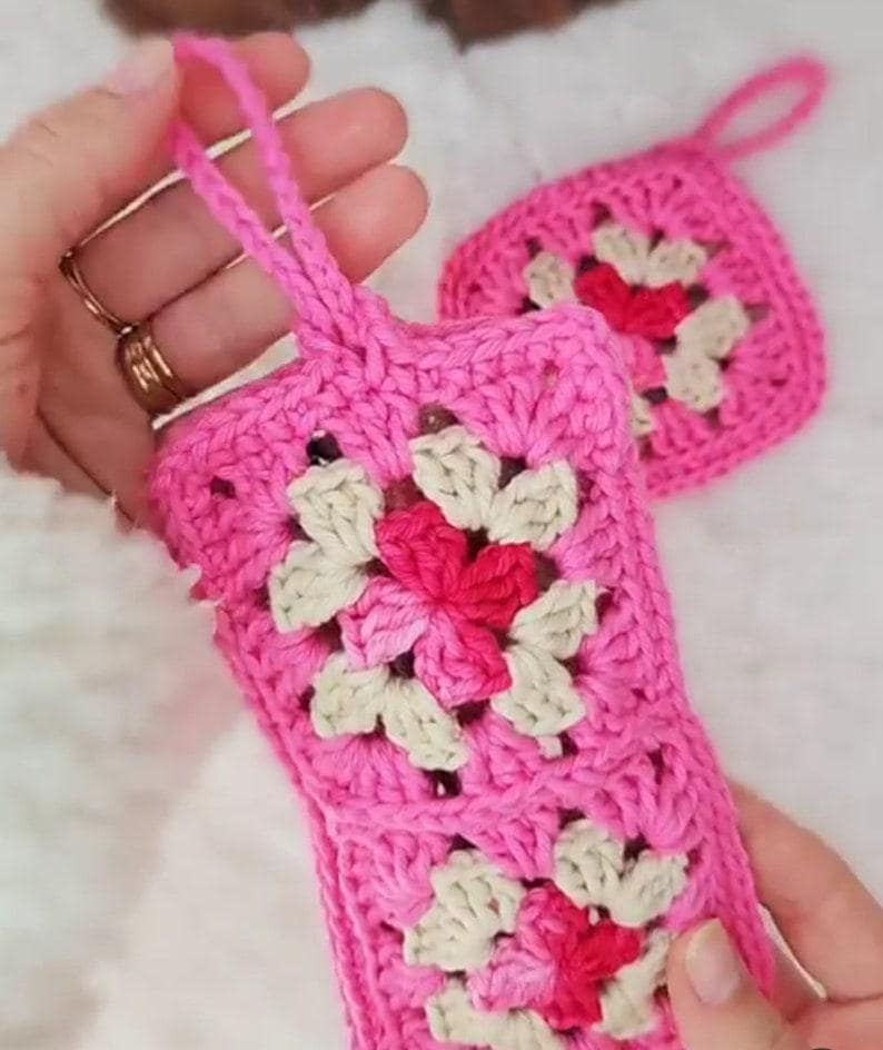 Op de afbeelding: Roze gehaakte telefoonhoes en bijpassende vierkante onderzetter. De hoes heeft een bloemenpatroon met witte en rode accenten. De hoes heeft een lus om te dragen. De onderzetter heeft een vergelijkbaar bloemenpatroon.