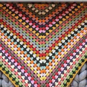 May include: A colorful, handmade crochet shawl. The shawl features a granny square design with a variety of colors, including red, orange, yellow, green, blue, and purple. The edges are finished with a yellow and green border. The shawl is displayed on a gray surface.