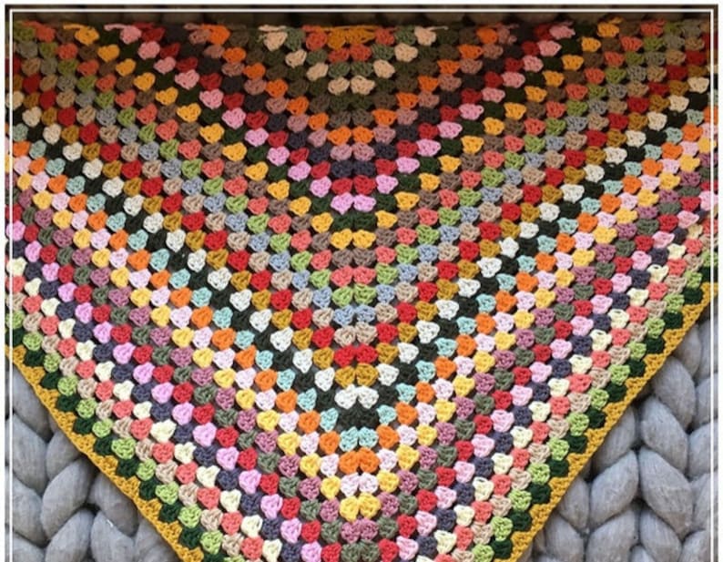 May include: A colorful, handmade crochet shawl. The shawl features a granny square design with a variety of colors, including red, orange, yellow, green, blue, and purple. The edges are finished with a yellow and green border. The shawl is displayed on a gray surface.