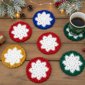 May include: A collection of handmade crochet coasters in various colors, including red, blue, green, and yellow. Each coaster features a white snowflake design. A green mug with coffee sits on a matching coaster, with holiday decorations in the background.