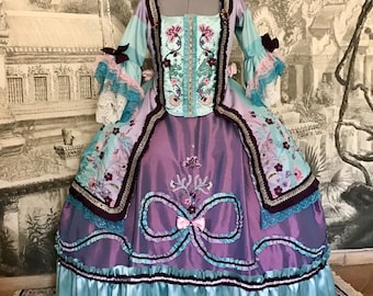 May include: A historical-style gown in shades of blue, purple, and burgundy, displayed on a mannequin. The bodice features floral embroidery and a square neckline. The skirt has a layered design with ribbon accents and a ruffled hem. The sleeves are long and flared.