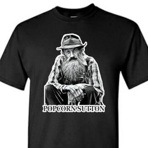 May include: Black t-shirt featuring a black and white illustration of a man with a long beard and hat. The text "POPCORNSUTTON" is printed below the illustration. The shirt is a crew neck style.