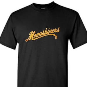 May include: Black t-shirt with the word "Moonshiners" in a gold, cursive font across the chest. The text has a slight shadow effect, adding depth to the design. The shirt is a classic crew neck style.