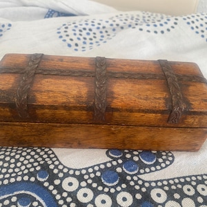 Può includere: Una scatola rettangolare in legno con finitura marrone medio, caratterizzata da fasce metalliche marrone scuro con una trama intrecciata che attraversano il coperchio. La scatola presenta una superficie liscia e lucida dal design semplice.