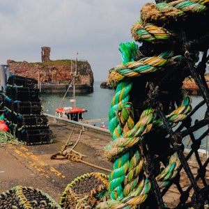 May include: A coastal scene featuring a harbor with fishing equipment. A close-up of a thick, multi-colored rope is in the foreground. In the background, there are lobster traps, a red and white boat, and a stone structure on a small island.