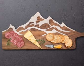 Puede incluir: Una tabla de cortar de madera con forma de cordillera montañosa con una incrustación de epoxi blanca. La tabla presenta una variedad de aperitivos, incluyendo salami, queso y galletas.