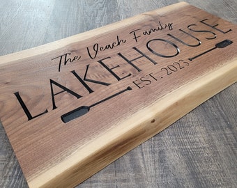 Puede incluir: Un letrero de madera con el texto "The Beach Family LAKEHOUSE EST. 2023" grabado en letras negras. El letrero presenta un diseño de remo estilizado.