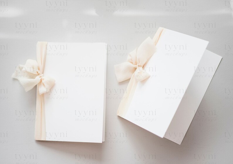 May include: Two blank white cards with a beige ribbon tied around one of the cards. The cards are on a light gray background.