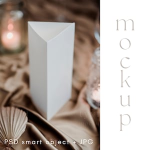 May include: A white triangular box sits on a brown fabric surface. The box is a mockup for a product or design. The text "MOCKUP" is written vertically on the right side of the image.