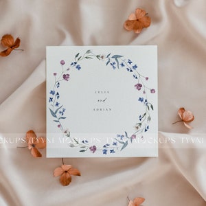 Pu&ograve; includere: Un biglietto bianco con un design a corona di fiori ad acquerello. La corona &egrave; composta da fiori blu e rosa e foglie verdi. Il testo "Celia e Adrian" &egrave; scritto al centro del biglietto.
