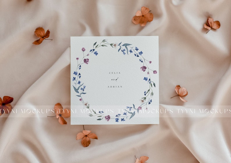 Pu&ograve; includere: Un biglietto bianco con un design a corona di fiori ad acquerello. La corona &egrave; composta da fiori blu e rosa e foglie verdi. Il testo "Celia e Adrian" &egrave; scritto al centro del biglietto.