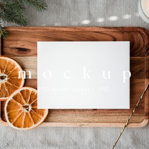 May include: A blank white card labeled "mockup" rests on a wooden tray, surrounded by dried orange slices and a sprig of evergreen. A lit candle and a gold bar tool are also present, creating a warm, inviting aesthetic. The image is a mockup for stationery or print design.