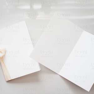May include: Two blank white cards with a light brown ribbon tied in a bow on one of the cards. The cards are on a light gray surface.