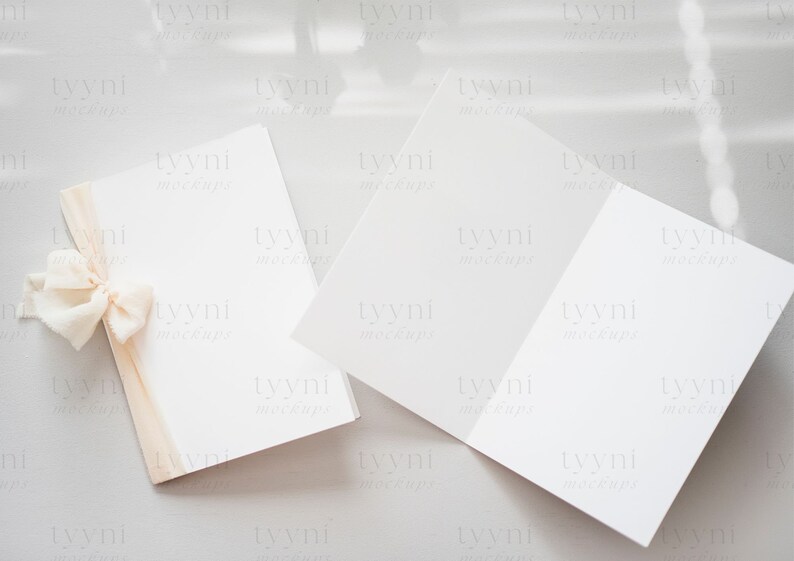 May include: Two blank white cards with a light brown ribbon tied in a bow on one of the cards. The cards are on a light gray surface.