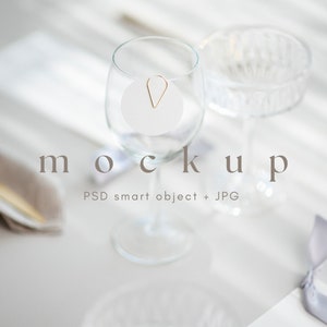 May include: A white round card with a gold clip attached to a clear glass wine glass. The glass is on a white table with a white napkin and a gold utensil. The text "mockup" is printed below the glass.