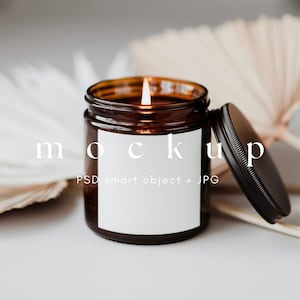 May include: A brown glass candle jar with a white label and a black lid. The candle is lit and the jar is sitting on a white surface with dried palm leaves.