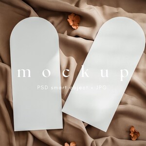 Peut inclure: Deux cartes de maquette blanches arquées avec le mot "mockup" imprimé dessus, reposant sur un tissu beige. Les cartes sont positionnées en biais, avec des fleurs séchées éparpillées autour. Le texte "PSD smart object + JPG" est également visible.
