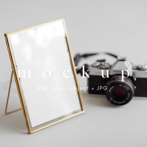 May include: A gold-toned metal picture frame with a white background, standing on a white surface. A vintage camera is out of focus in the background. The text "mockup PSD smart object + JPG" is printed below the frame.