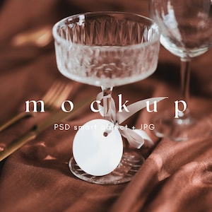 May include: A white round tag with a hole in the center, tied with a white ribbon, sits on a clear glass goblet. The goblet is on a brown fabric surface with gold cutlery.