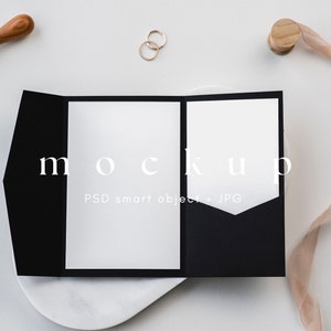 May include: A black invitation mockup with white inserts, featuring the word "mockup". Accompanying the invitation are gold rings, a wooden stamp, wax seals, and a ribbon. The design is set against a white background.