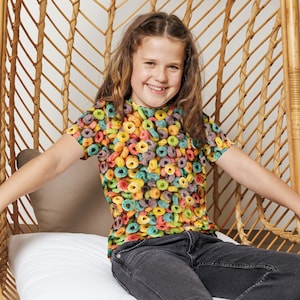 May include: A young person wearing a colorful t-shirt with a cereal print sits in a wicker chair. The chair has a white cushion and the person is wearing dark jeans and pink sneakers.