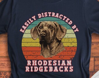 Puede incluir: Una camiseta azul oscuro con un gráfico de estilo vintage de un perro Rhodesian Ridgeback. El gráfico presenta un fondo de puesta de sol con el texto "Easily Distracted By Rhodesian Ridgebacks".
