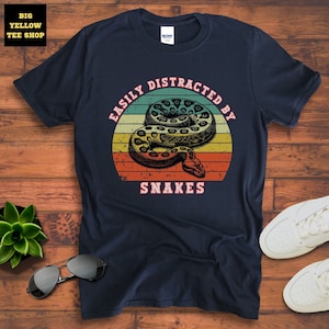 Op de afbeelding: Een marineblauw t-shirt met een vintage-stijl afbeelding van een slang met de tekst "Easily Distracted By Snakes".