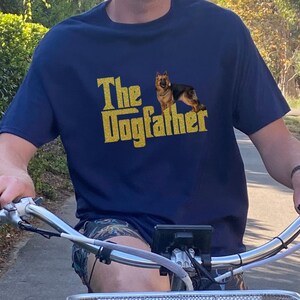 May include: A person wearing a navy blue t-shirt with a gold glitter design that says "The Dogfather" and a German Shepherd silhouette. The person is riding a bicycle.