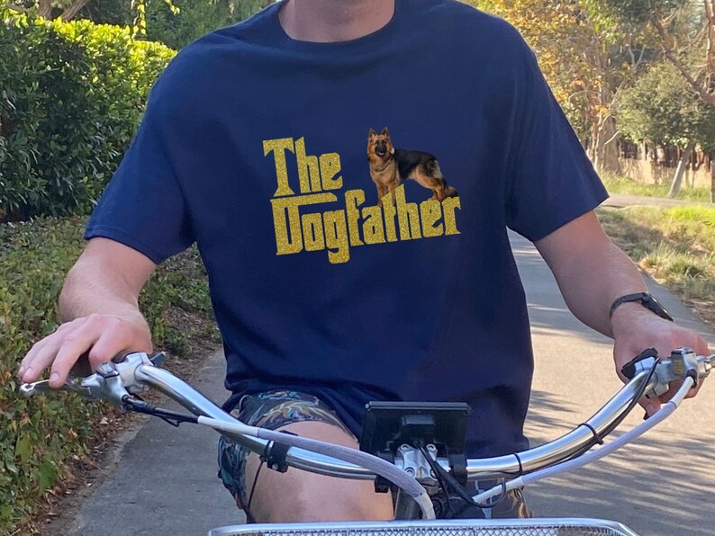 May include: A person wearing a navy blue t-shirt with a gold glitter design that says "The Dogfather" and a German Shepherd silhouette. The person is riding a bicycle.