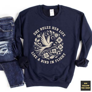 May include: Navy blue sweatshirt with the text "SHE RULES HER LIFE LIKE A BIRD IN FLIGHT" in a circular design featuring a bird and floral elements. The sweatshirt is paired with blue jeans.