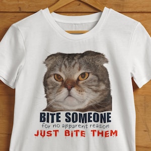 May include: White t-shirt featuring a close-up of a grumpy-looking cat with yellow eyes. Below the cat is the text "BITE SOMEONE for no apparent reason JUST BITE THEM" in black and red font.