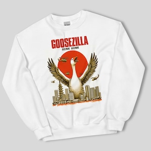 Può includere: Felpa bianca a maniche lunghe con una stampa grafica di un'oca con le ali spiegate, in piedi su uno skyline urbano con un sole rosso alle sue spalle. Il testo "GOOSEZILLA HONK HONK" è stampato sopra l'oca.