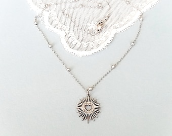 May include: A silver necklace with a delicate chain and small beads. The pendant is a silver sunburst with a heart-shaped cutout in the centre.