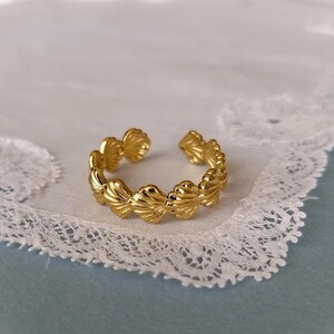 May include: A gold-colored ring with a seashell design. The ring is open at the top and features a series of detailed seashell shapes. The ring is set against a white lace background.