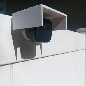 Peut inclure: Un support de caméra de sécurité rectangulaire blanc avec une caméra de sécurité noire attachée. Le support est monté sur une clôture blanche. La caméra est noire et possède un objectif. Le support protège la caméra du soleil.