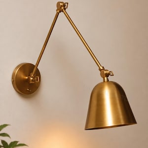 May include: A brass wall-mounted lamp featuring an adjustable swing arm with two articulated joints and a dome-shaped shade. The fixture has a circular mounting plate and a metallic finish, designed to extend outward from the wall surface.