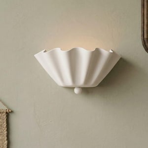 May include: A matte white wall sconce featuring a semi-circular, ruffled, shell-like design with a small spherical accent at the bottom centre. The fixture emits a soft, warm glow from its top opening, casting light upward against the wall surface.
