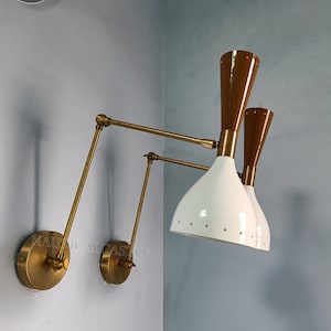 Może przedstawiać: Para lamp ściennych z mosiądzu z białymi i brązowymi kloszami. Lampy mają regulowane ramiona i są zamontowane na jasnoniebieskiej ścianie.