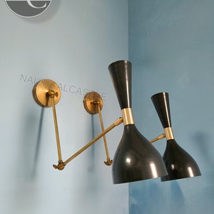 May include: Two black and gold sconce lights fixed to a light blue wall. The lights feature a cone-shaped shade and a brass arm. The brand name "Nautical Castle" is visible in the upper left corner.