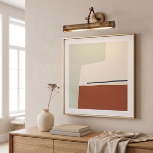 May include: An abstract art print in a gold frame hangs above a wooden cabinet. The artwork features geometric shapes in muted colors of rust, cream, and sage green. A vintage-style brass picture light is mounted above the artwork.