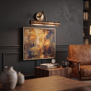 May include: An abstract painting with gold frame hangs on a dark wall, illuminated by a bronze-toned picture light. A wooden side table holds books, a clock, and a mug. A leather armchair is in the background.