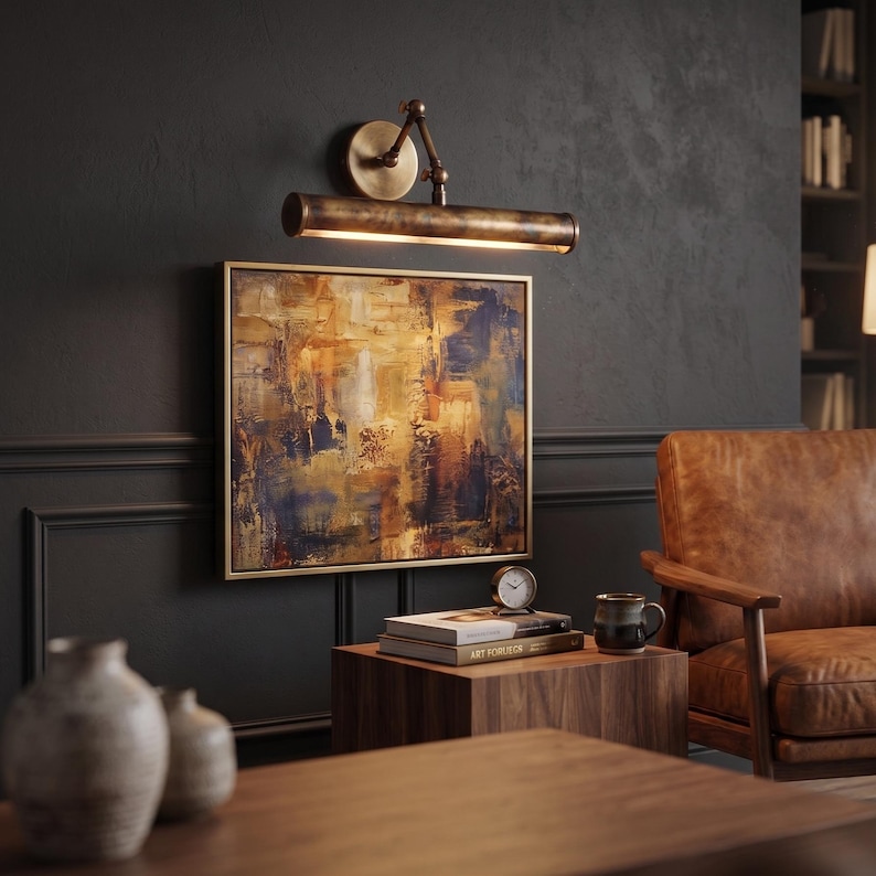 May include: An abstract painting with gold frame hangs on a dark wall, illuminated by a bronze-toned picture light. A wooden side table holds books, a clock, and a mug. A leather armchair is in the background.