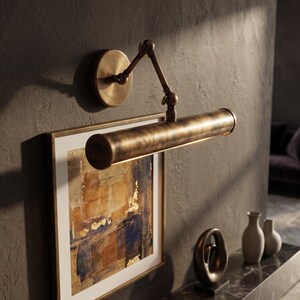 May include: A bronze-toned picture light is mounted on a textured gray wall, illuminating a framed abstract painting. The light has an adjustable arm and a cylindrical shade. Decorative vases and a sculpture are on a nearby surface.