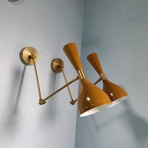 Puede incluir: Un par de apliques de pared de latón con brazos ajustables y pantallas de color amarillo dorado. Las pantallas tienen un interior blanco y un patrón de pequeños agujeros.