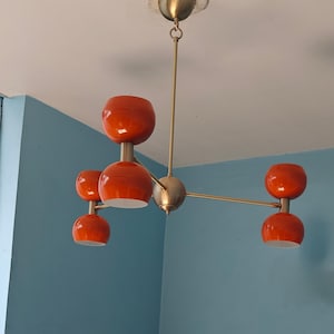 May include: A mid-century modern chandelier with five orange glass shades. The fixture has a brass finish and is suspended from the ceiling. The shades are a vibrant orange color, and the fixture is a stylish lighting option.