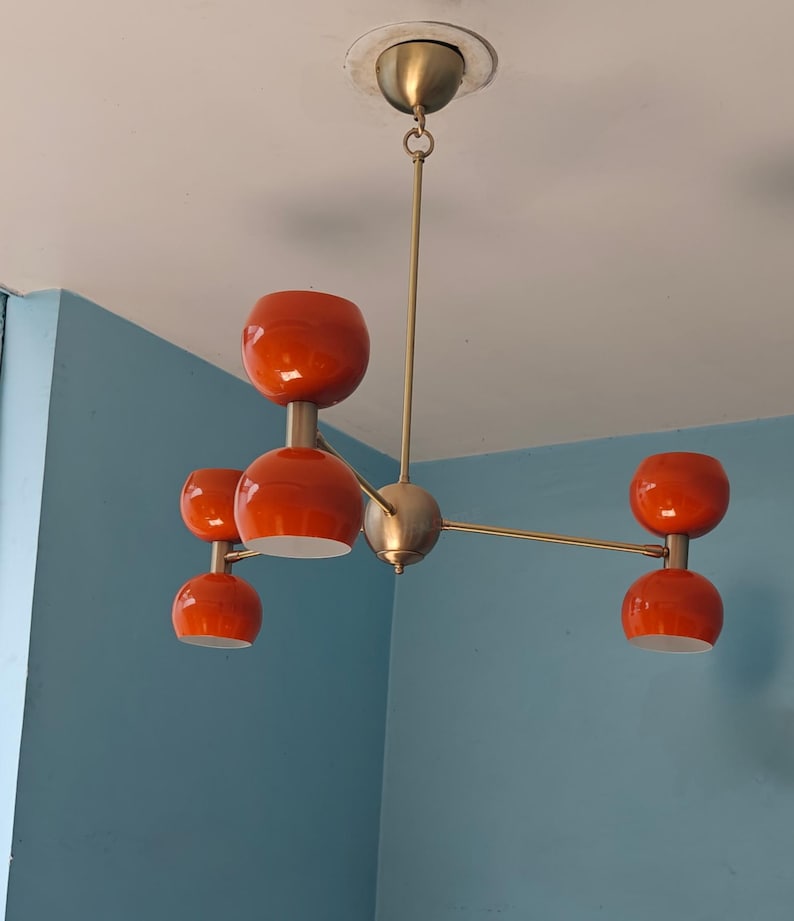 May include: A mid-century modern chandelier with five orange glass shades. The fixture has a brass finish and is suspended from the ceiling. The shades are a vibrant orange color, and the fixture is a stylish lighting option.