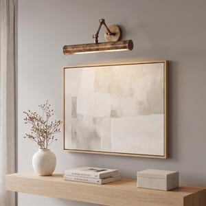 May include: A bronze-toned picture light is mounted above a framed abstract painting. A white vase with dried branches, two books, and a small box sit on a wooden shelf. The painting has a neutral color palette.